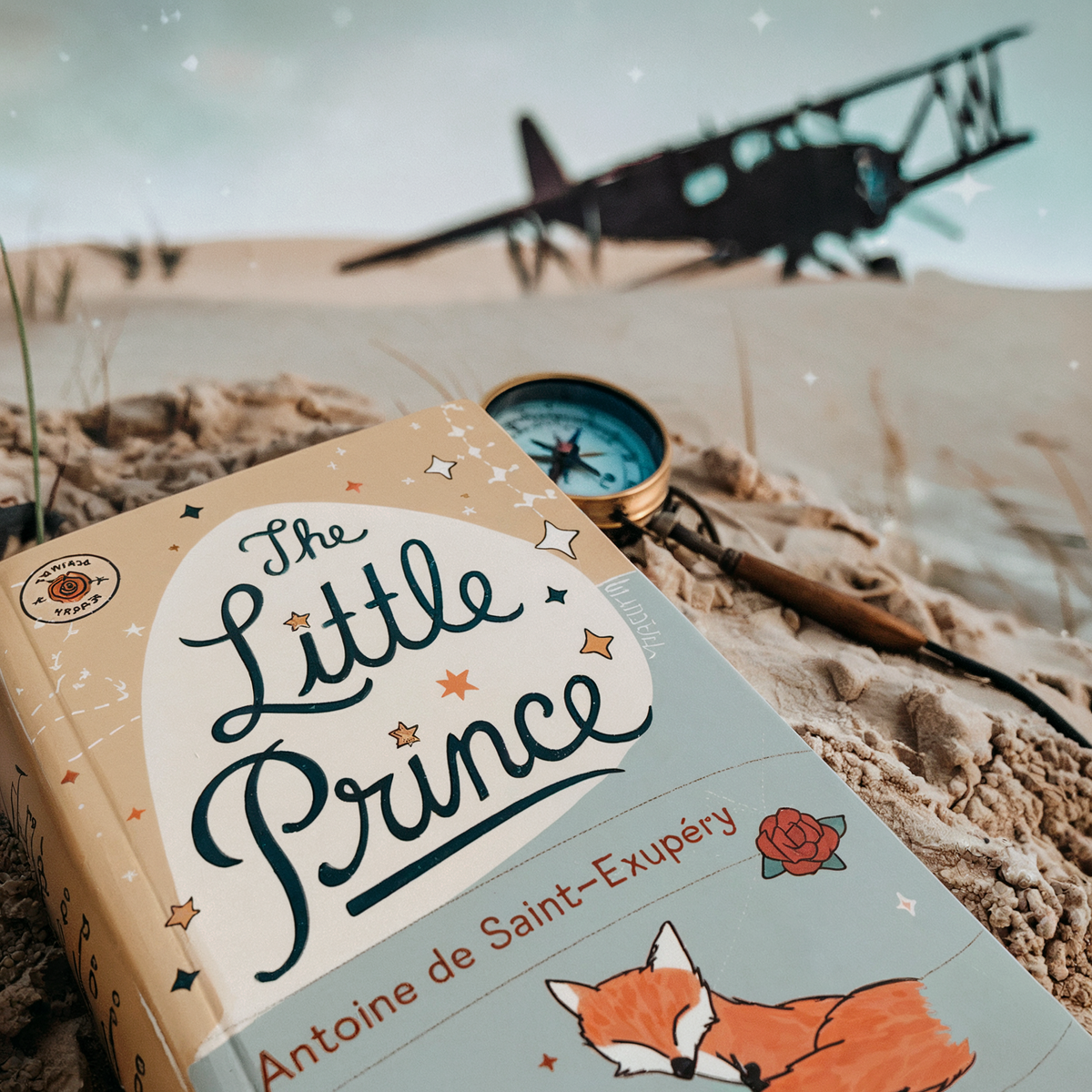 "The Little Prince" book by Antoine de Saint-Exupéry on sand with a compass and an airplane in the background.